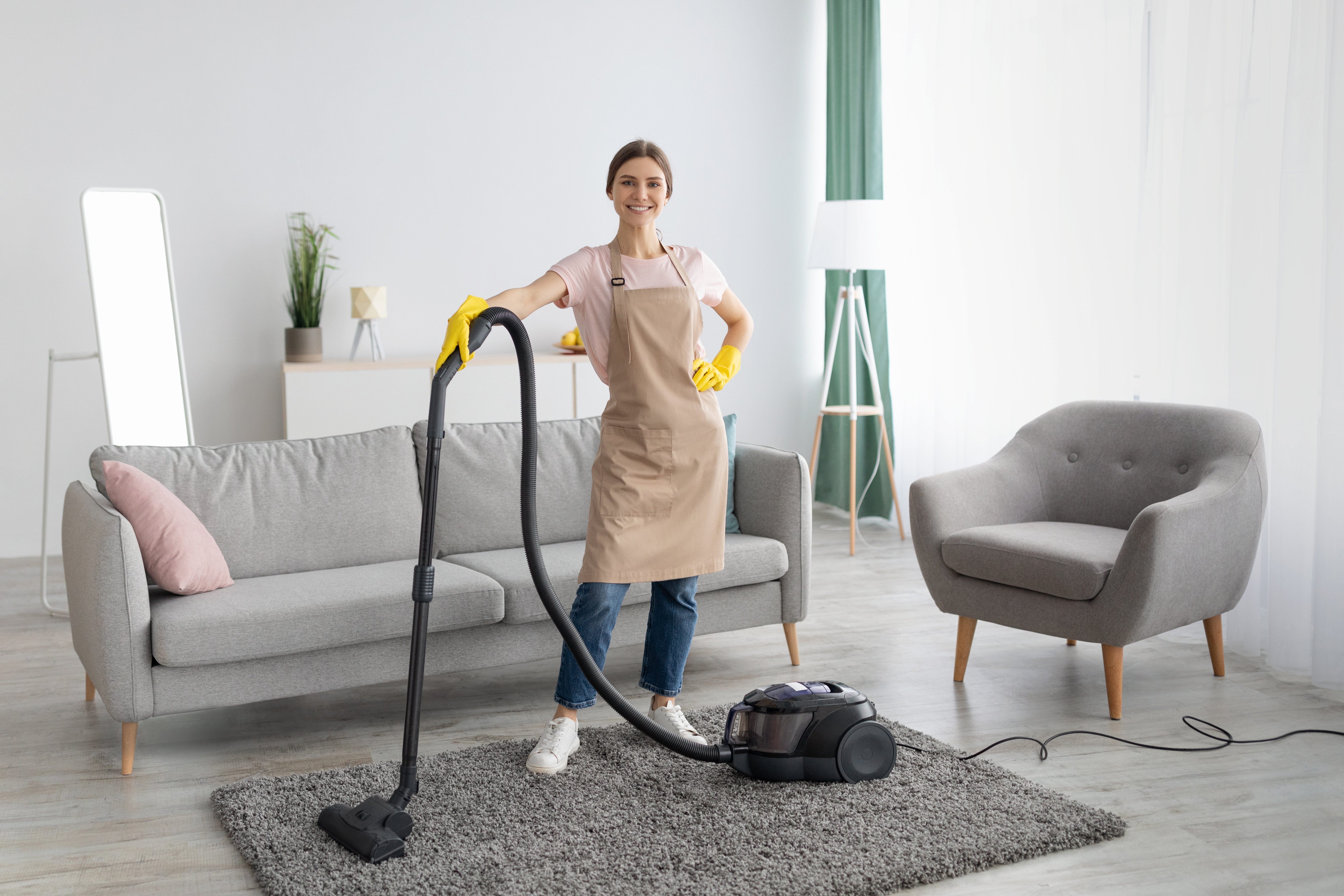 Professional cleaning service. Positive young lady in apron and rubber gloves vacuuming living room of modern house